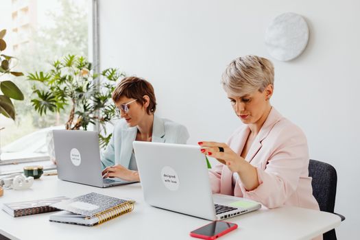 Two businesswomen working collaboratively on laptops in a bright, modern office setting with natural light.
