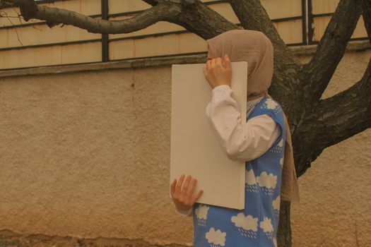 A woman wearing a hijab holds a blank whiteboard against a textured wall outside.
