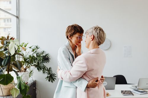 Free Two women embracing lovingly in a modern office filled with natural light and greenery. Stock Photo