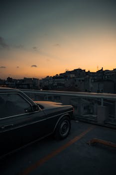 Old car parked on rooftop with cityscape silhouette against a sunset sky, capturing a serene urban evening.
