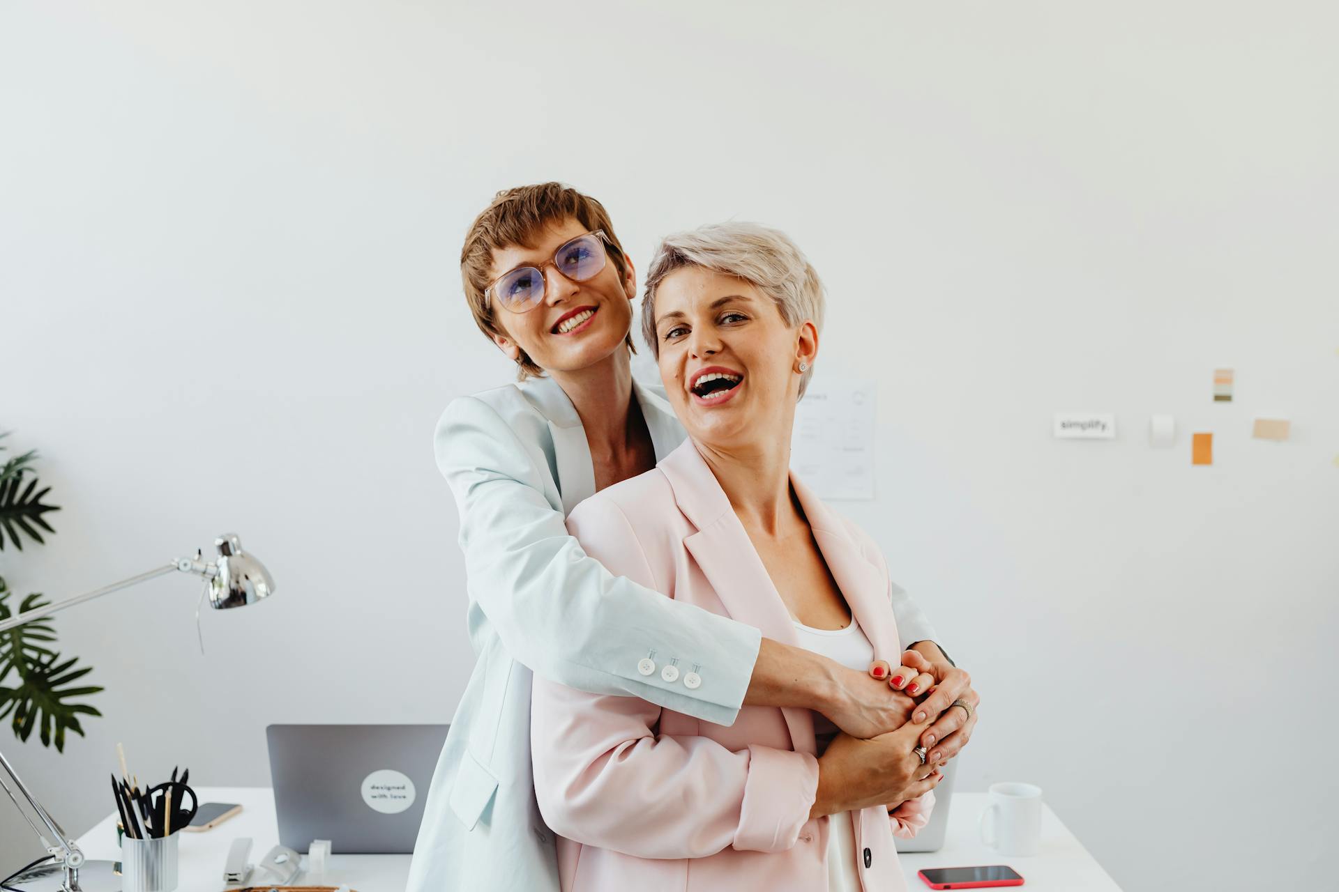 Two businesswomen in corporate attire sharing a joyful embrace in a modern office setting.