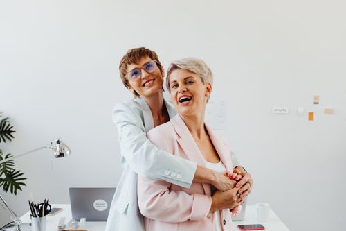 Free Two businesswomen in corporate attire sharing a joyful embrace in a modern office setting. Stock Photo