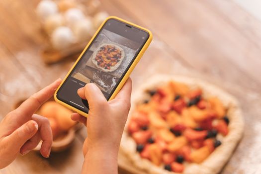 Hands taking a photo of a homemade fruit pie with a smartphone in a warm kitchen setting.