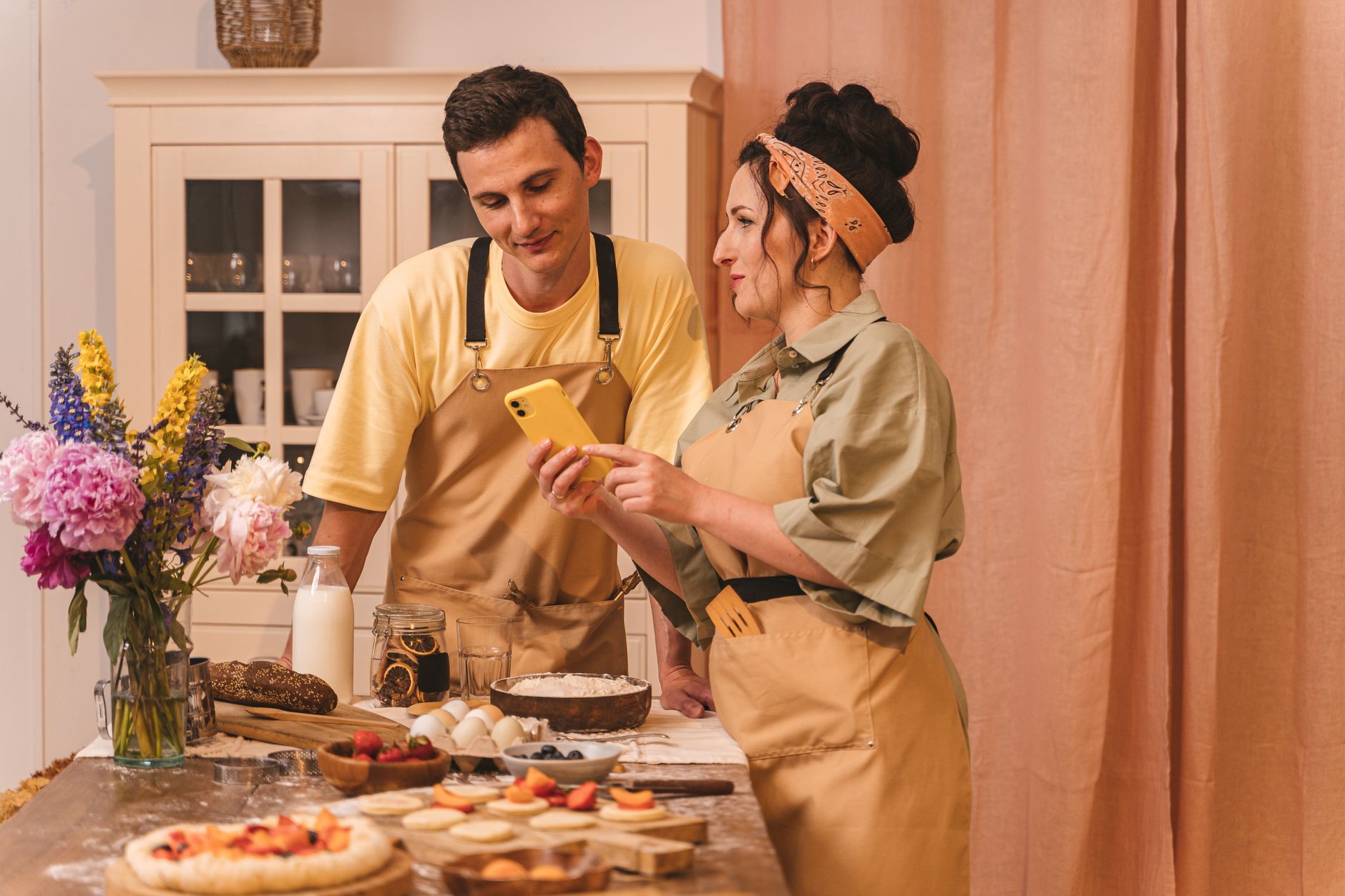 A couple in aprons preparing food and using a smartphone in a cozy kitchen setting.