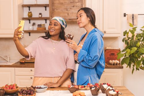 Free Two women capturing a joyful moment while enjoying desserts and taking selfies in a cozy kitchen setting. Stock Photo