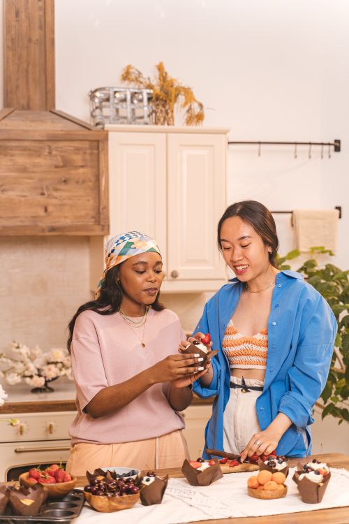 Free Two women enjoying a fun baking session in a cozy kitchen, decorating homemade cupcakes with colorful toppings. Stock Photo