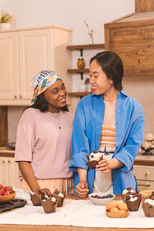 Free Two friends smiling and enjoying cupcakes together in a cozy kitchen setting. Stock Photo