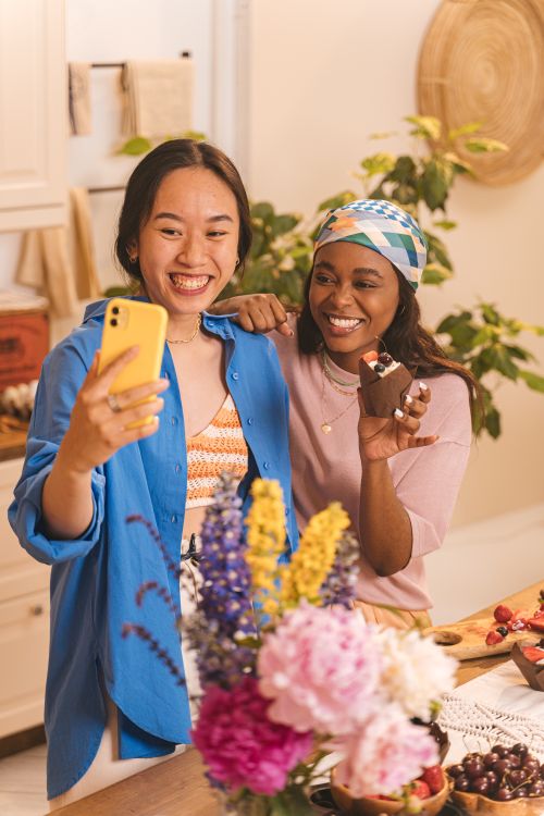 Free Two friends indoors smiling, enjoying cupcakes and taking a selfie together in a cozy setting. Stock Photo