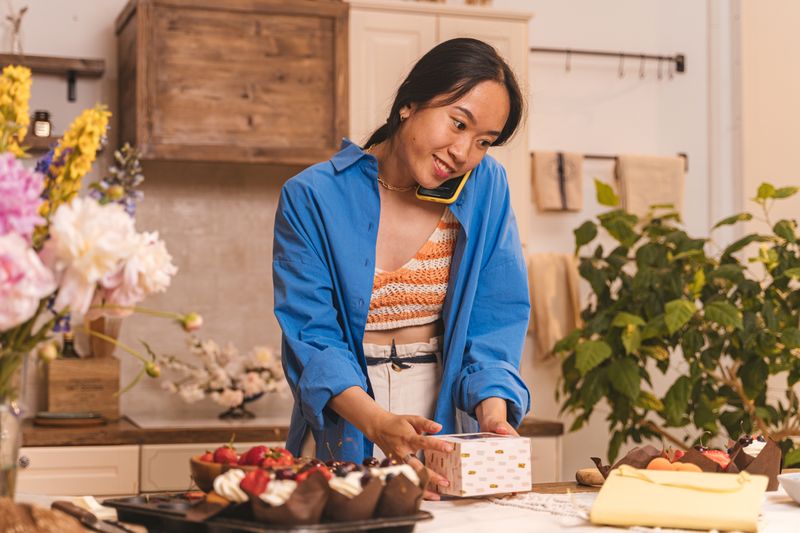 Bakery owner smiling on a phone call while packing boxes of cupcakes