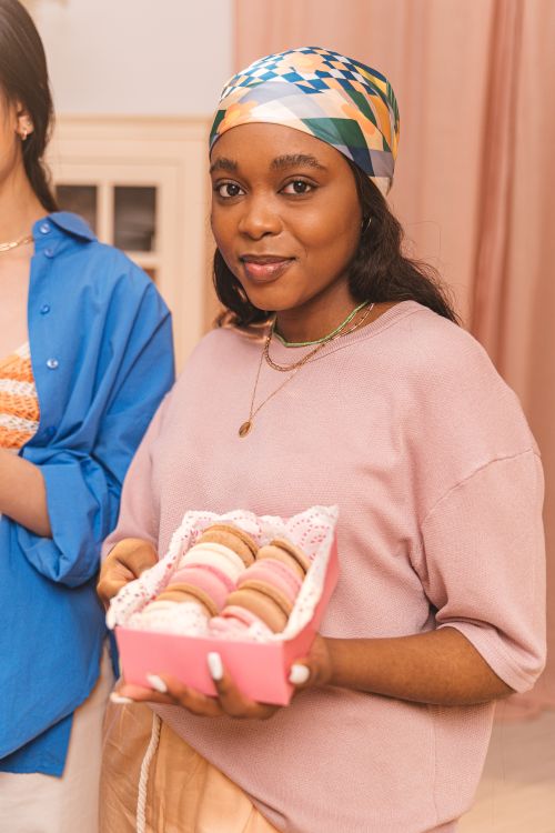 Free A woman in a cozy home setting holding a box of colorful macarons, looking at the camera. Stock Photo