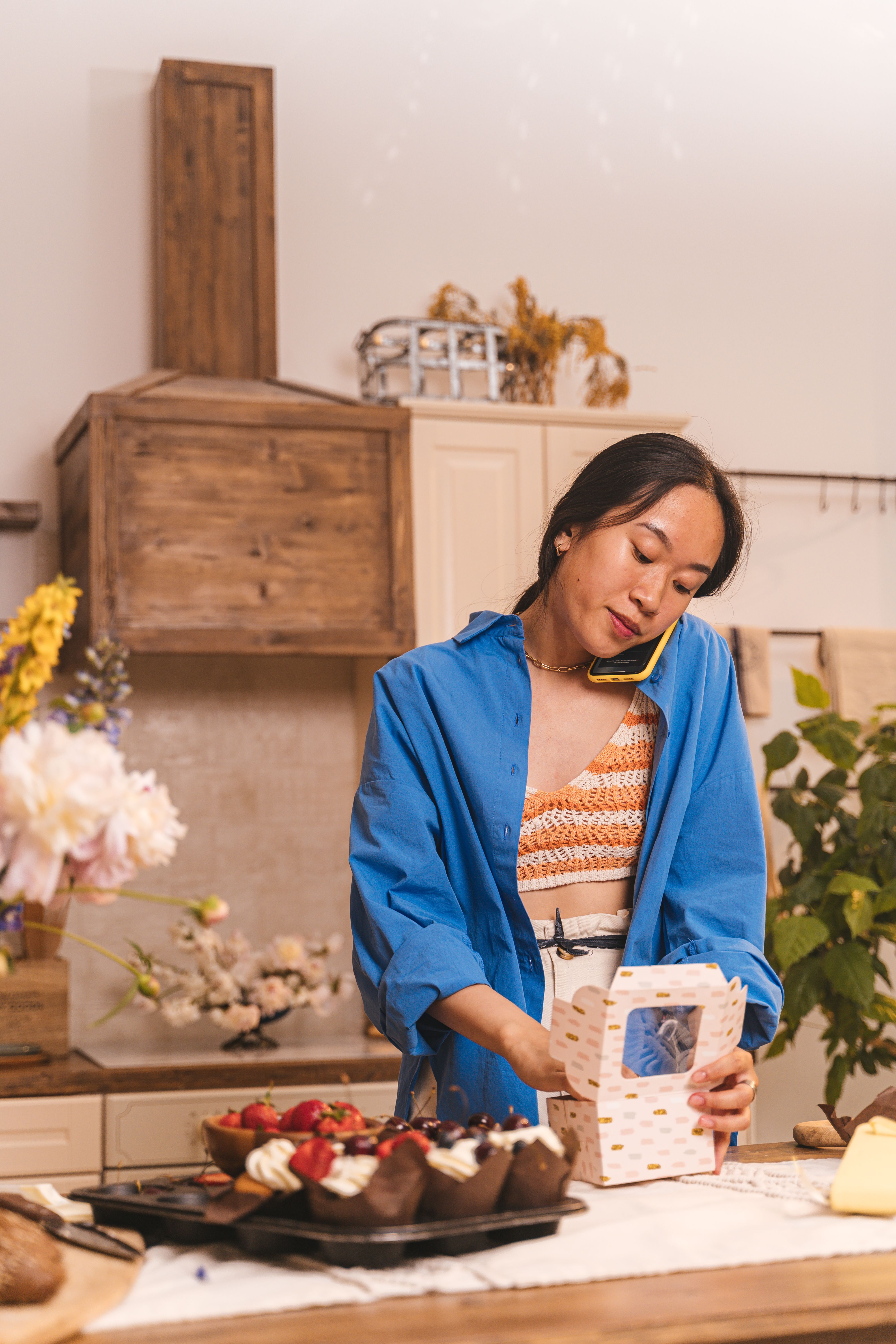 Asian woman multitasking by baking pastries and talking on the phone in a cozy kitchen setting.