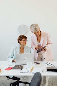Two women in elegant suits collaborating in a bright office setting.