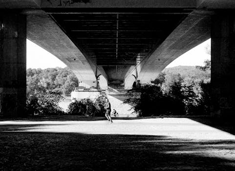A person jogging under a large bridge in a dramatic grayscale setting.