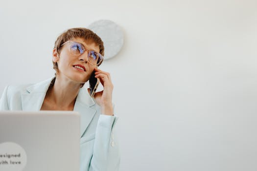 Woman in office setting, wearing glasses, talking on smartphone, and exuding professionalism.