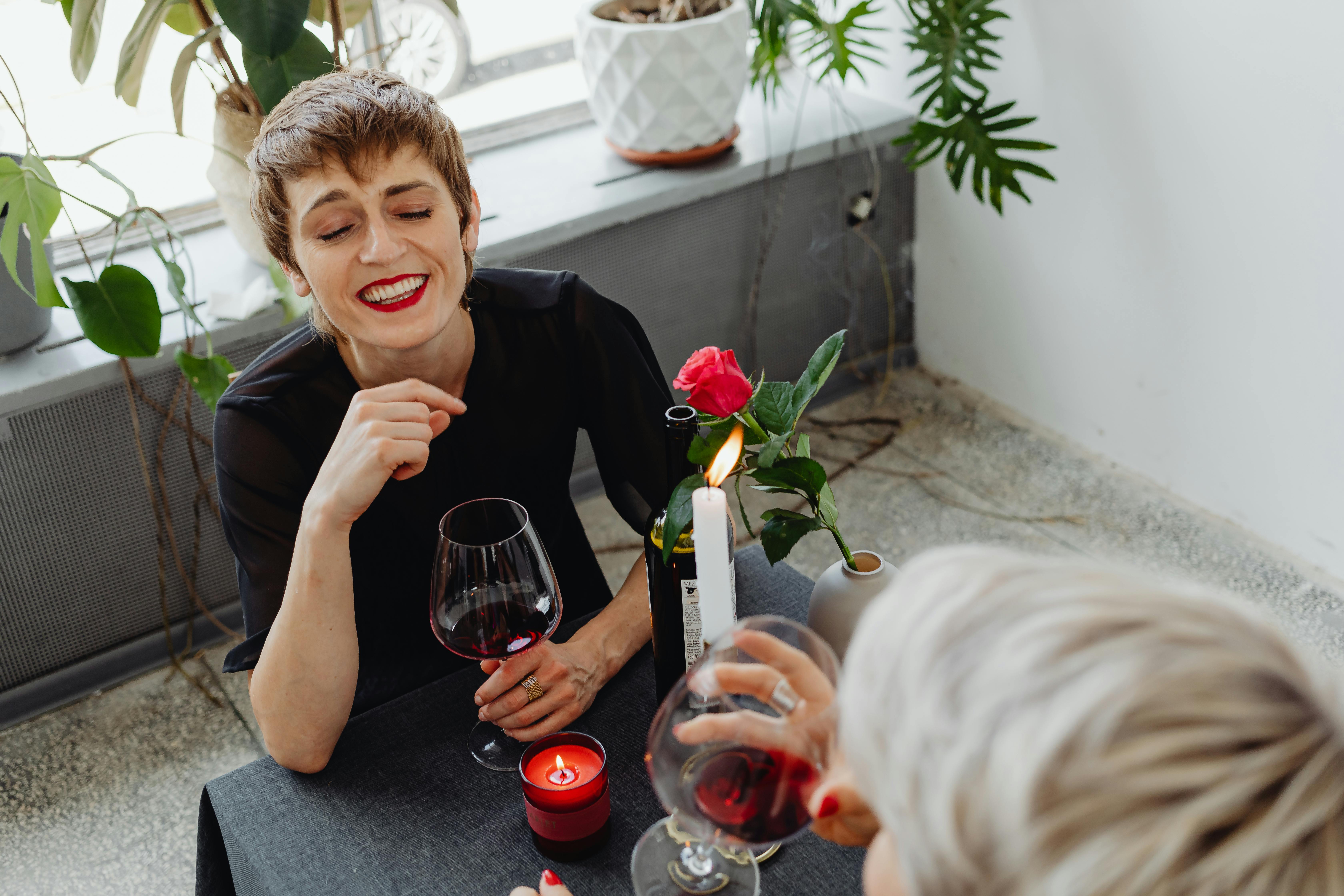 Two women enjoying a cozy, romantic dinner indoors, laughing over wine with candles and flowers.