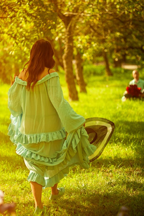 Woman in Green Dress with Sun Hat Walking on Grass Field