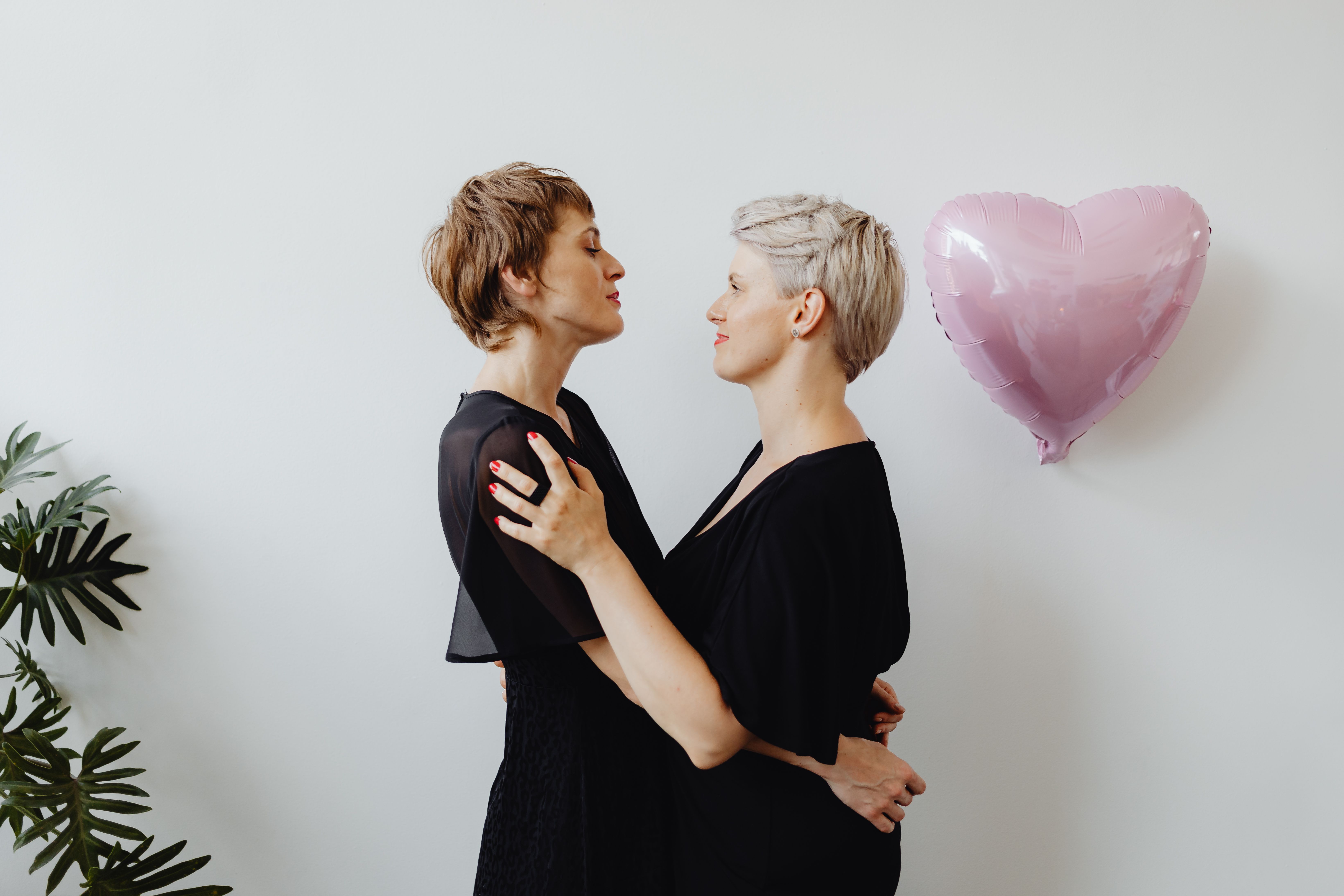 A loving lesbian couple embraces indoors with a pink heart-shaped balloon symbolizing lgbt pride.