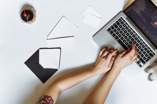 Top view of a person typing on a laptop with envelopes and a cupcake on a white desk.
