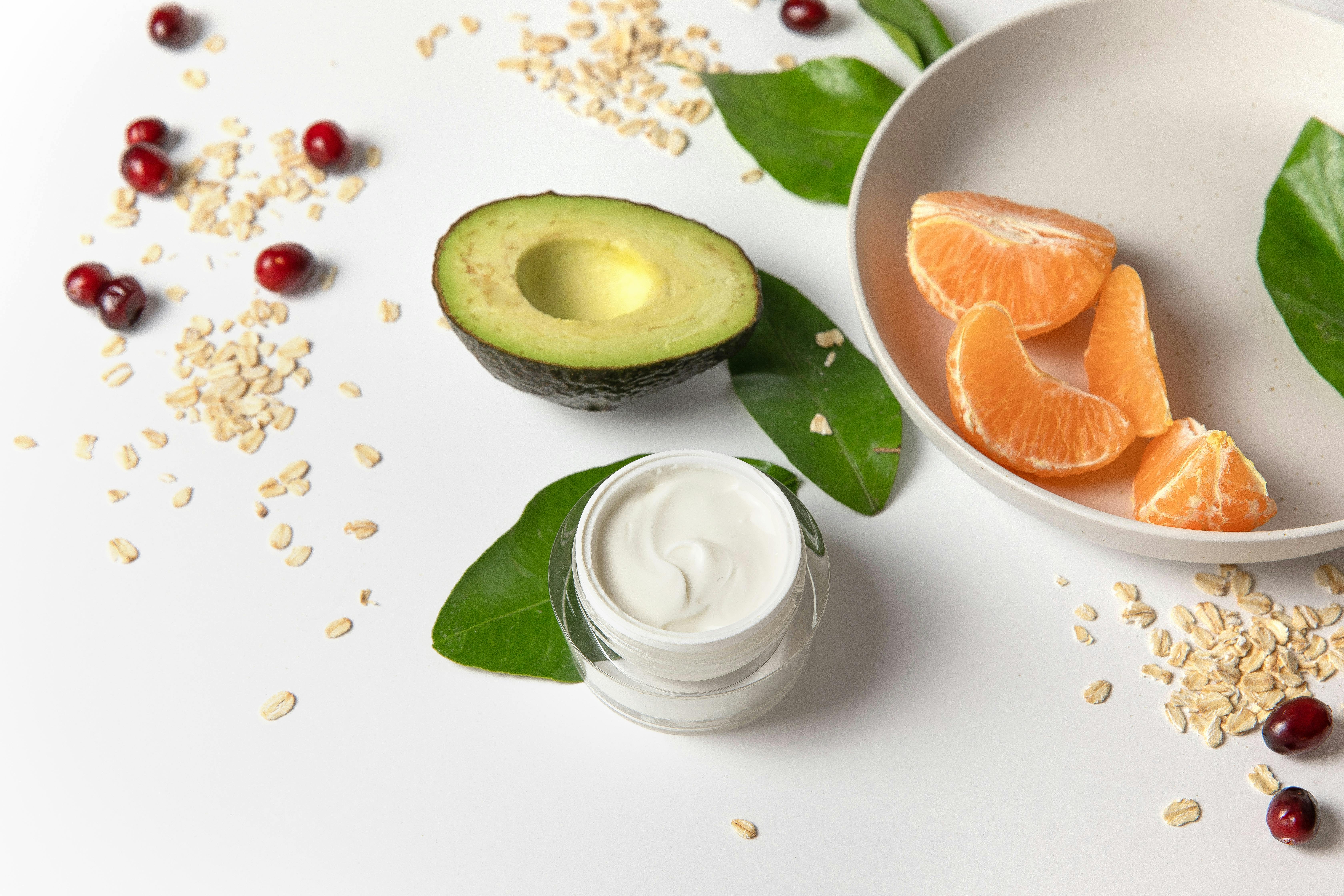 Fresh avocado, cream, and fruits on a white surface for skincare and healthy eating.