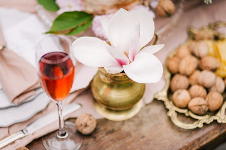 White Moth Orchids In Golden Vase Beside A Clear Glass Of Wine And Platter Of Nuts