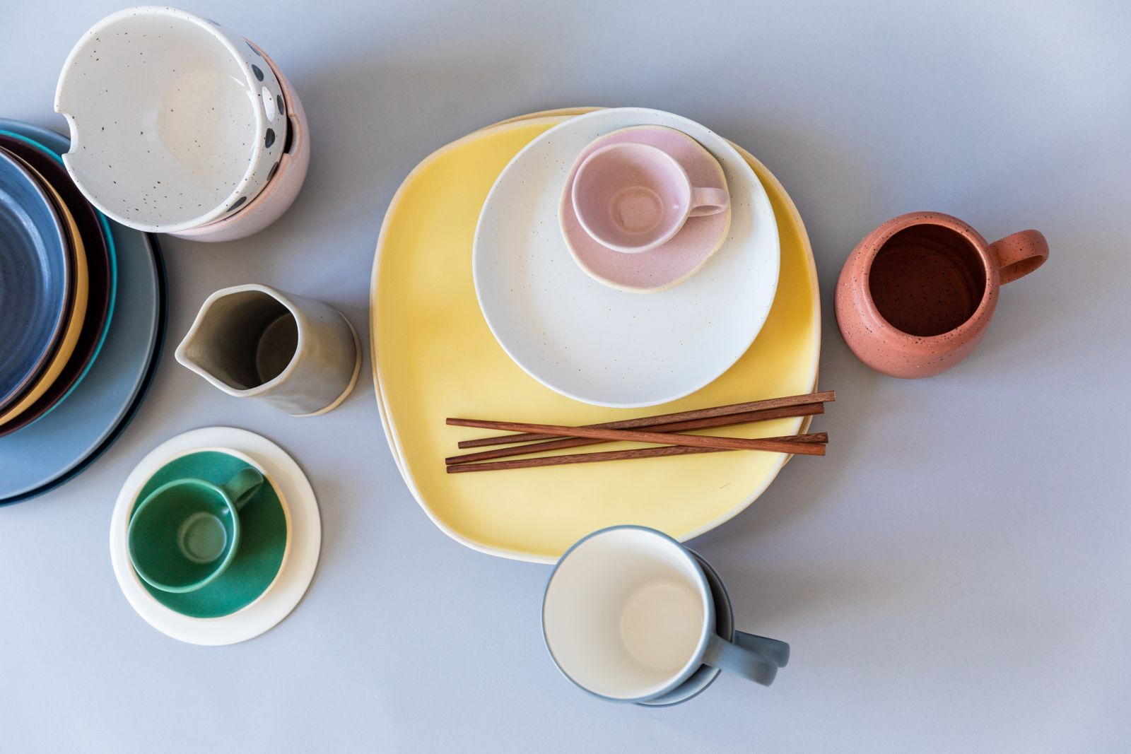 A flat lay of colorful ceramic tableware including plates, mugs, and chopsticks on a gray surface.