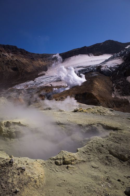 Free A captivating view of a volcano with smoke rising, surrounded by rugged terrain and glaciers under a blue sky. Stock Photo