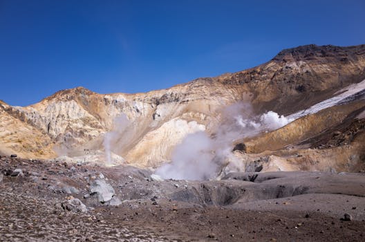 Explore the rugged beauty of a volcanic crater, complete with steaming geysers and striking rock formations under a clear blue sky.