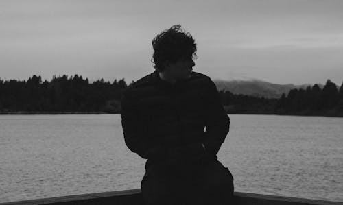 Grayscale Photo of a Man Sitting Near the Lake