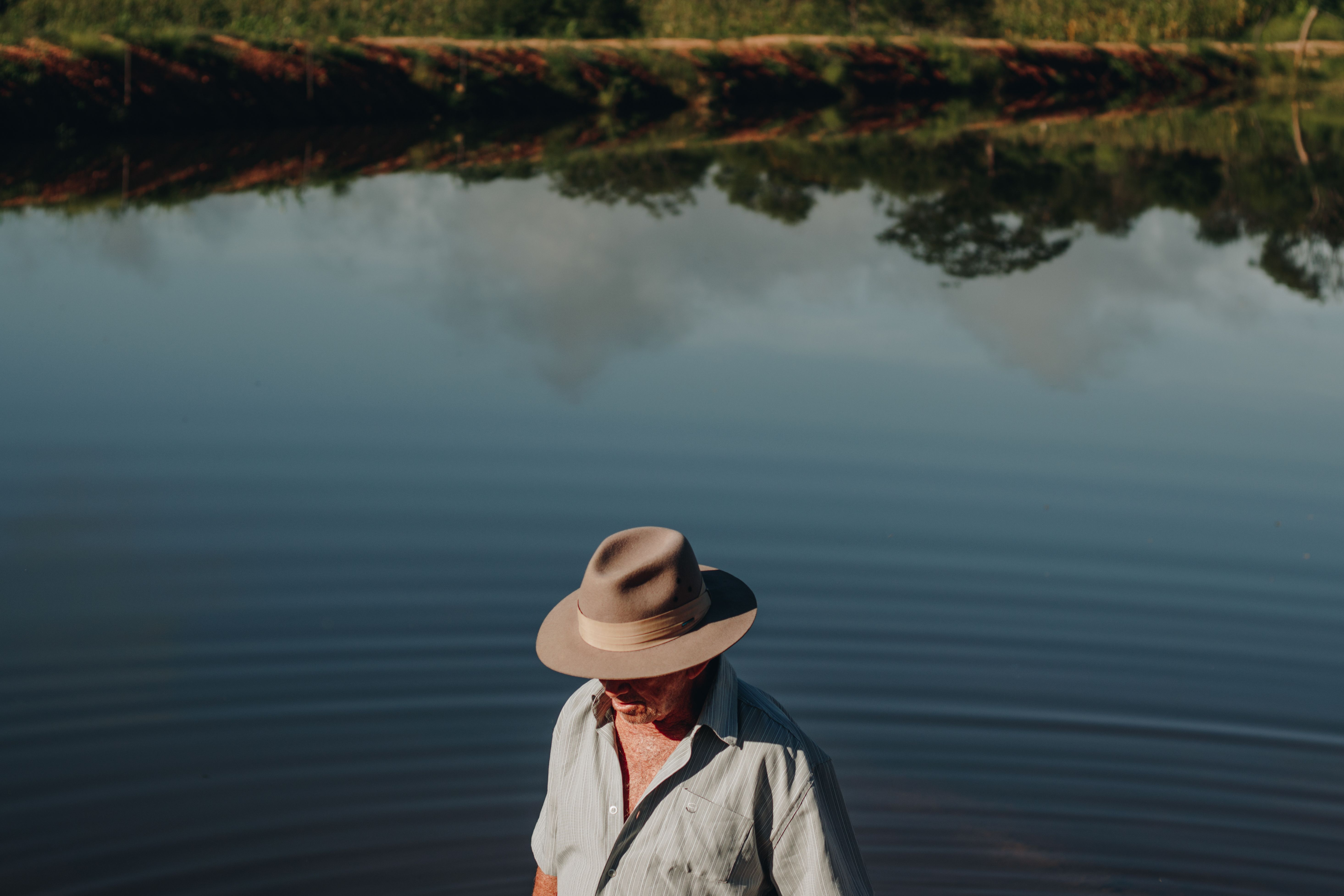 A man wearing a hat relaxes by a calm lake with trees reflecting in the water.
