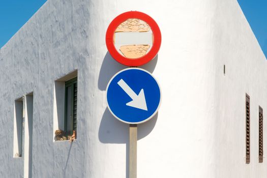 Blue and red road signs against a bright white wall under clear blue sky. Minimalist composition.