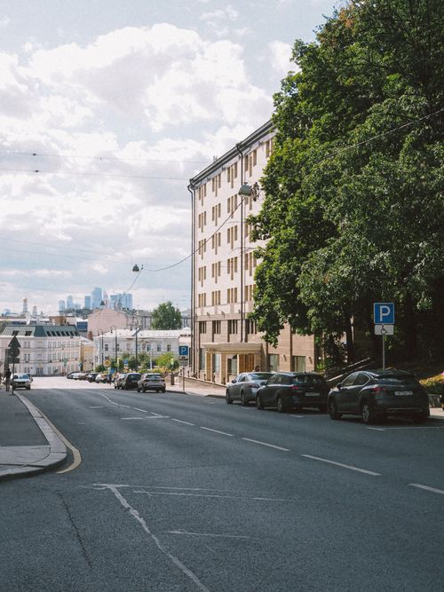 Free Street view of parked cars and urban buildings with trees lining the road under a cloudy sky. Stock Photo