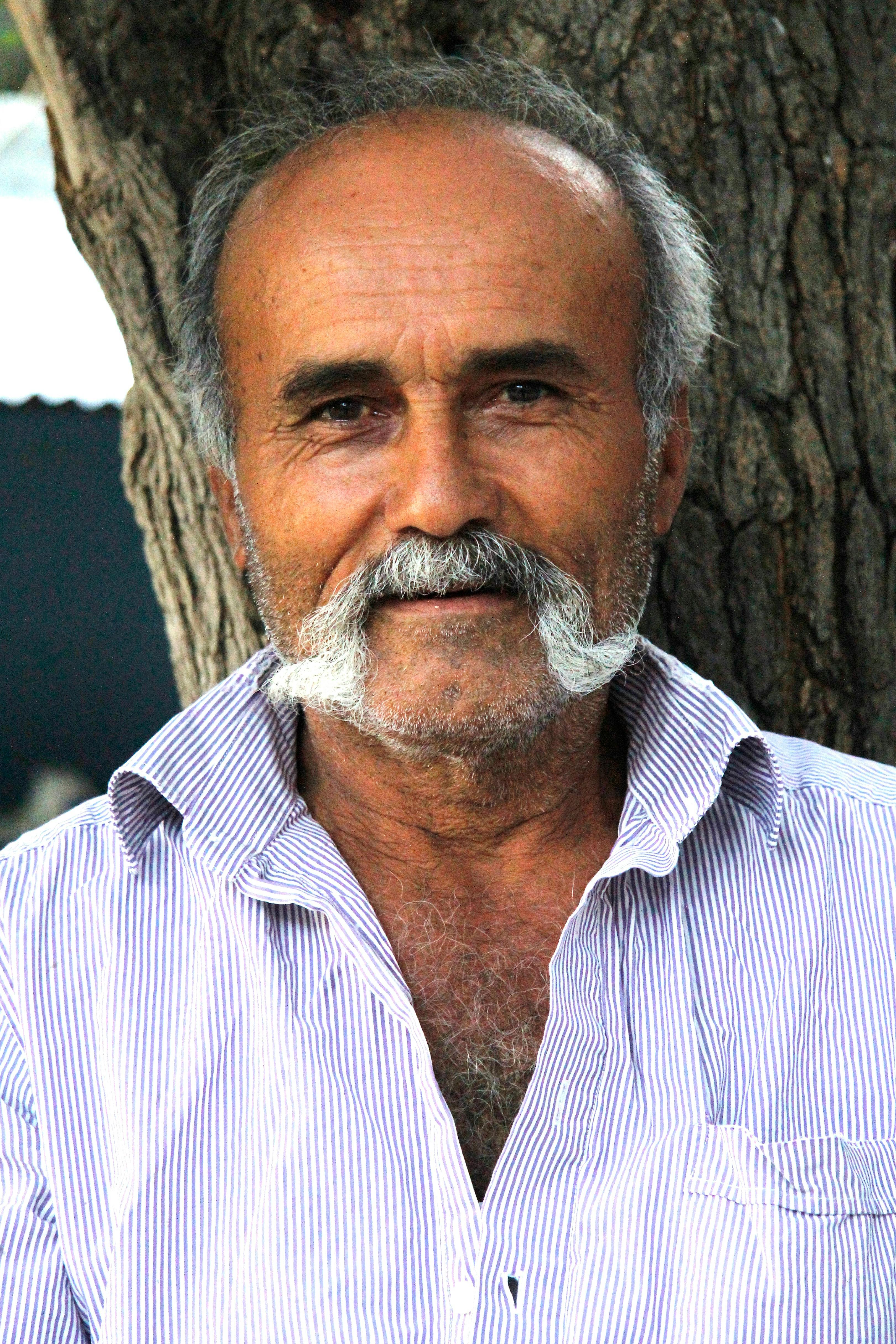 Portrait of a senior man with a bold mustache smiling warmly against a tree backdrop.