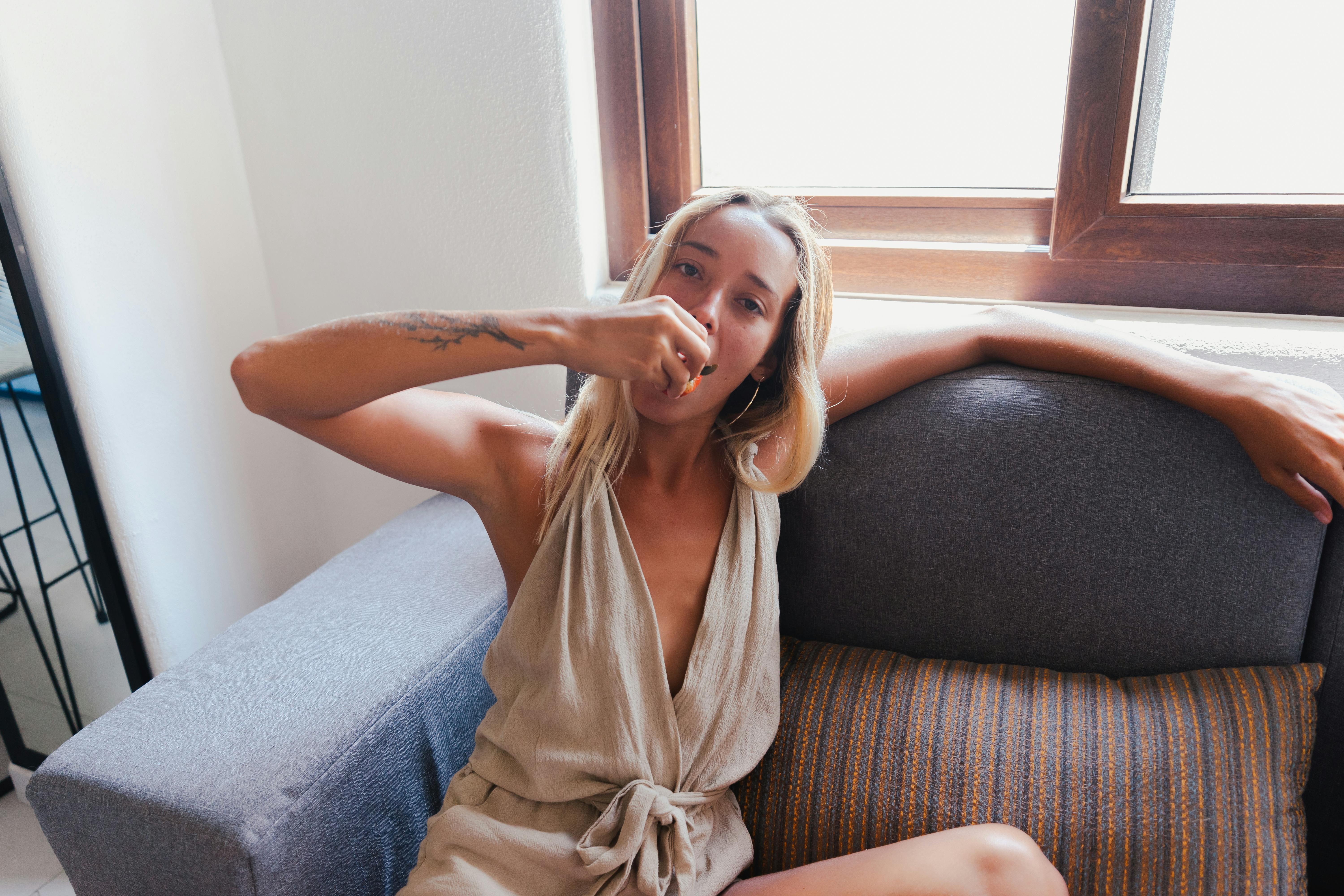 A woman sits comfortably on a couch near a window, enjoying a snack indoors.
