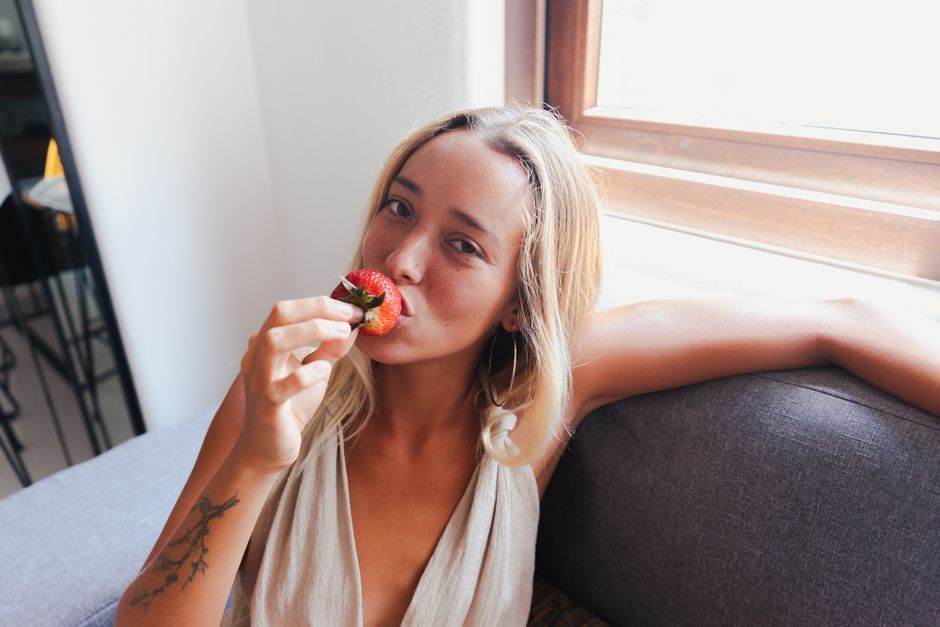 Blonde woman relaxing on a couch indoors, enjoys a fresh strawberry in natural light.