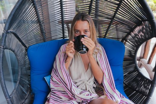 A woman enjoys a cozy drink on an outdoor swing chair wrapped in a blanket.