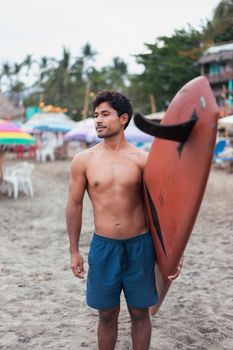 A man stands shirtless holding a surfboard on a tropical beach, ready for surf adventure.