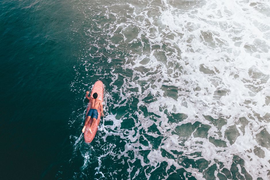 Aerial shot of a surfer paddling on a longboard in the ocean, surrounded by foamy waves.