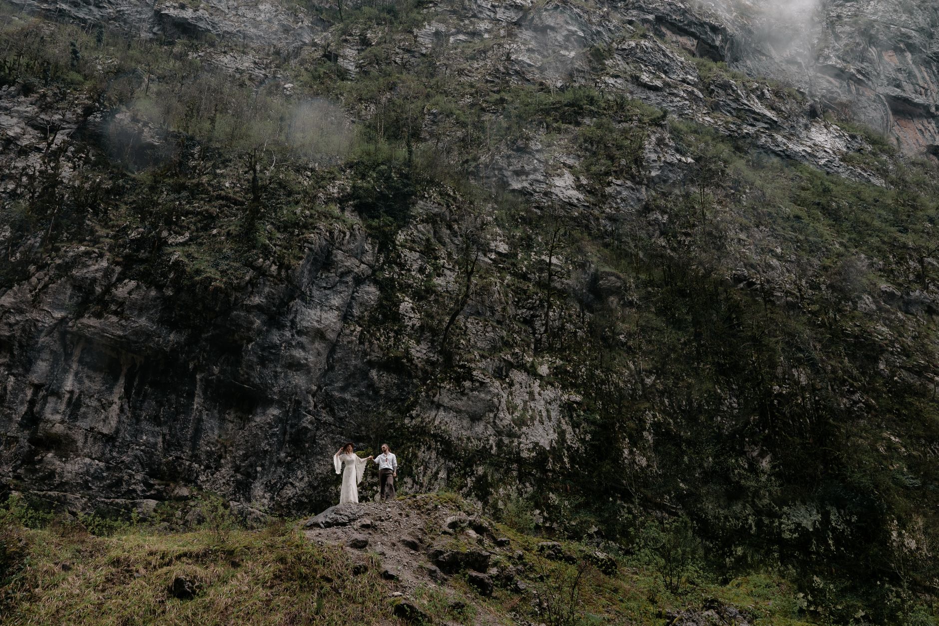 Couple dressed in formal attire stands against a towering rocky mountainside, embracing nature's grandeur.