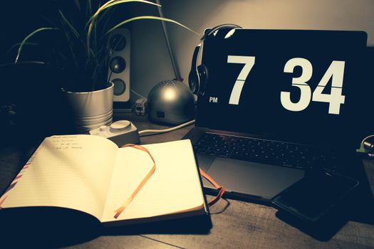 A cozy workspace with a laptop displaying time, open notebook, and headphones under warm lighting.