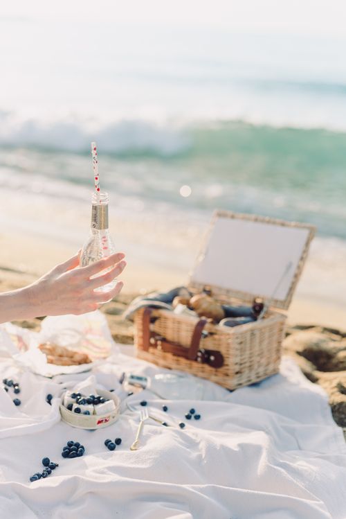 Free A serene beach picnic scene with a hand holding a glass bottle and a picnic basket. Stock Photo