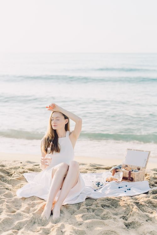 Free Woman in a white dress enjoying a peaceful beach picnic, sitting on sand by the sea. Stock Photo