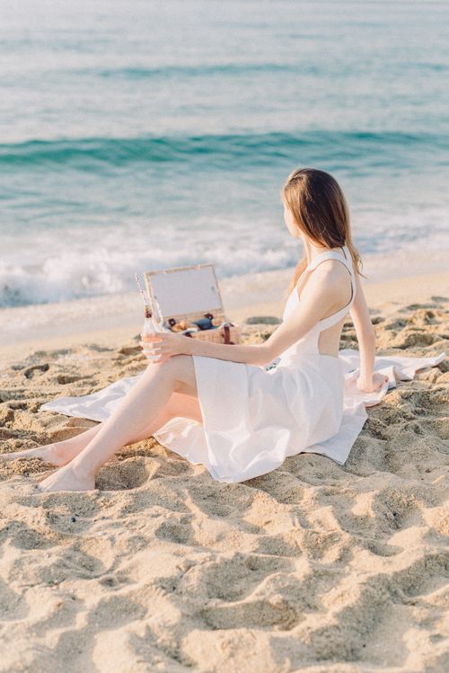 Free A woman in a white dress sitting on a sandy beach, gazing at the ocean waves on a sunny day. Stock Photo