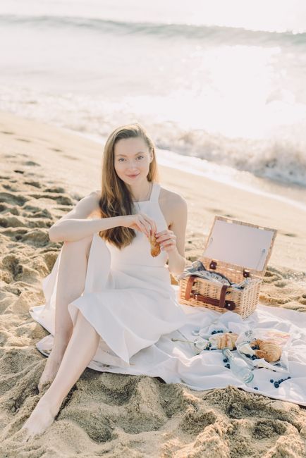 A woman enjoying a beach picnic with croissants, sitting by the ocean in a white dress.