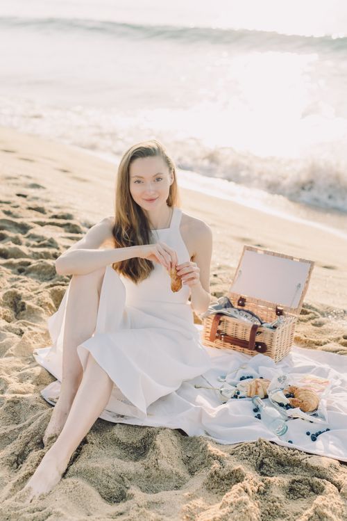 Free A woman enjoying a beach picnic with croissants, sitting by the ocean in a white dress. Stock Photo