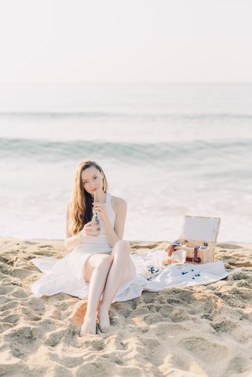 Free Young woman relaxes at a beach picnic sipping a beverage, surrounded by sand and sea. Stock Photo