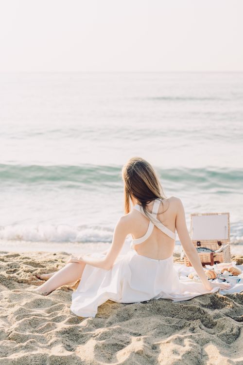 Free A woman in a white dress enjoys a serene beach picnic with a view of the gentle waves. Stock Photo