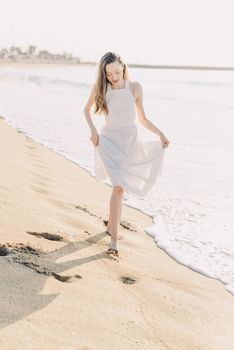 A woman in a white dress walking along a sunny beach with waves and footprints.