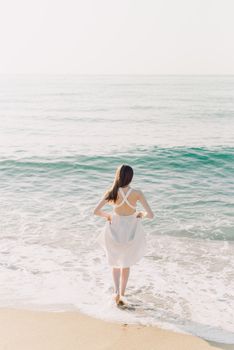 A woman in a white dress walks barefoot along a serene beach shoreline.