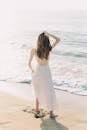 一名身穿白色连衣裙的女子站在阳光明媚的海滩上的背影，海浪拍打着海岸