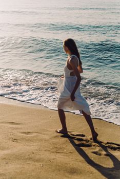 Woman in white dress walking on sandy beach with waves at sunset.